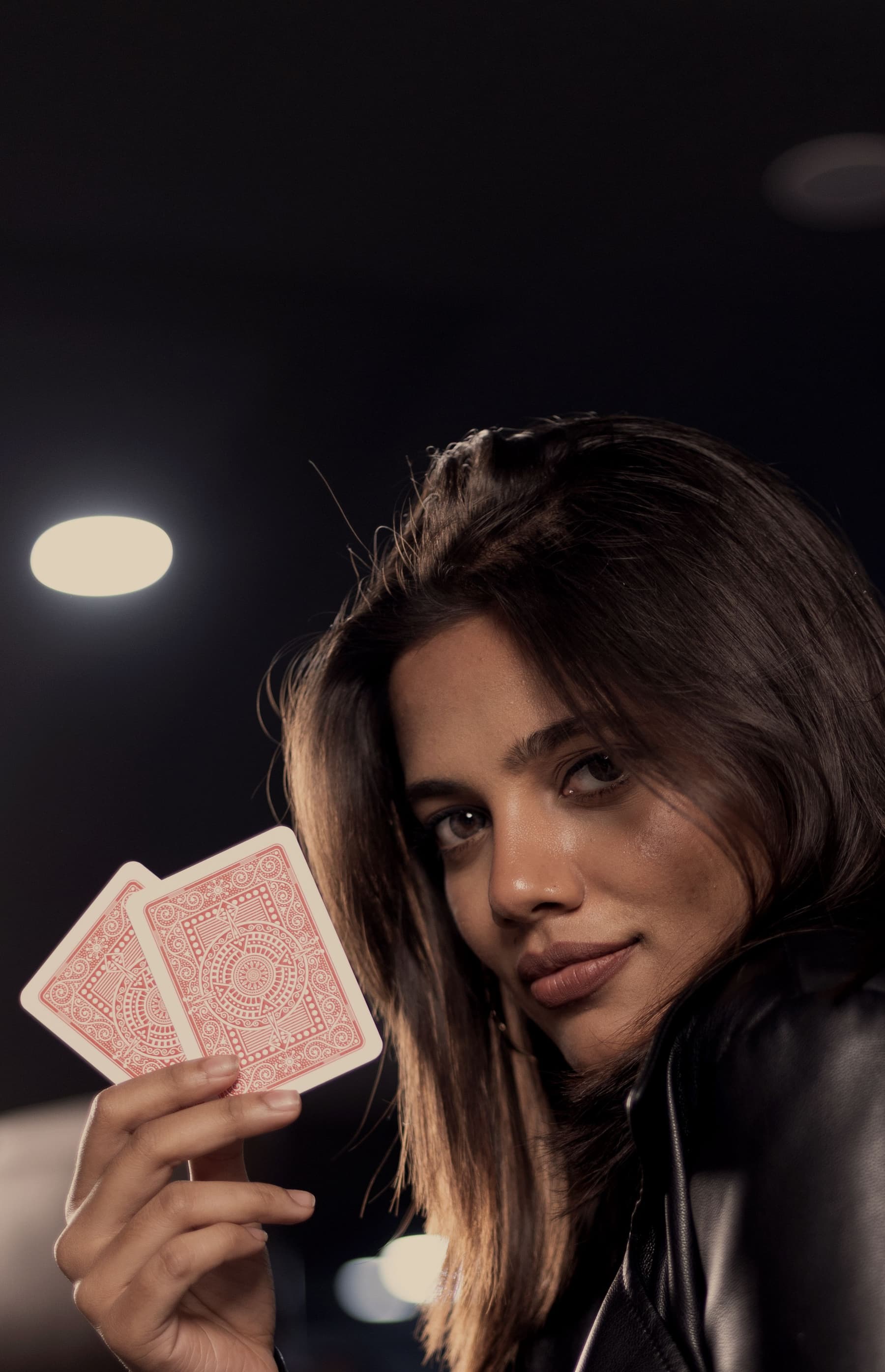 A player holding two cards at the table, the room’s ceiling lights behind her.
